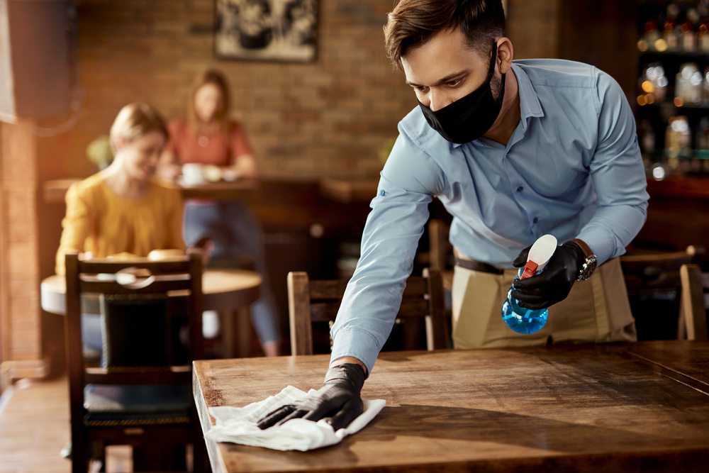 homem de mascara higienizando uma mesa num restaurante