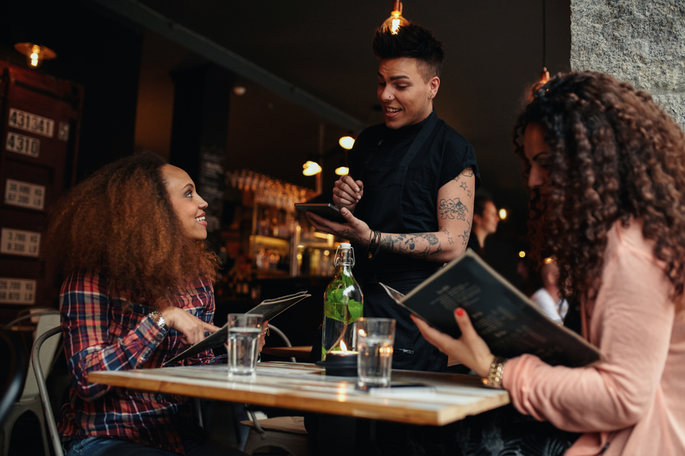 Mulheres olhando um cardápio numa mesa de bar enquanto atendente sorri
