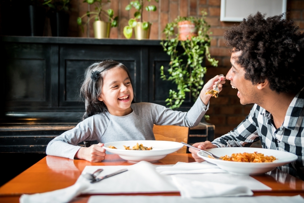 Homem preto, de camisa xadrez, sorri enquanto sua filha, uma criança, branca, de cabelos lisos, vestindo cinza, o alimenta com macarrão.