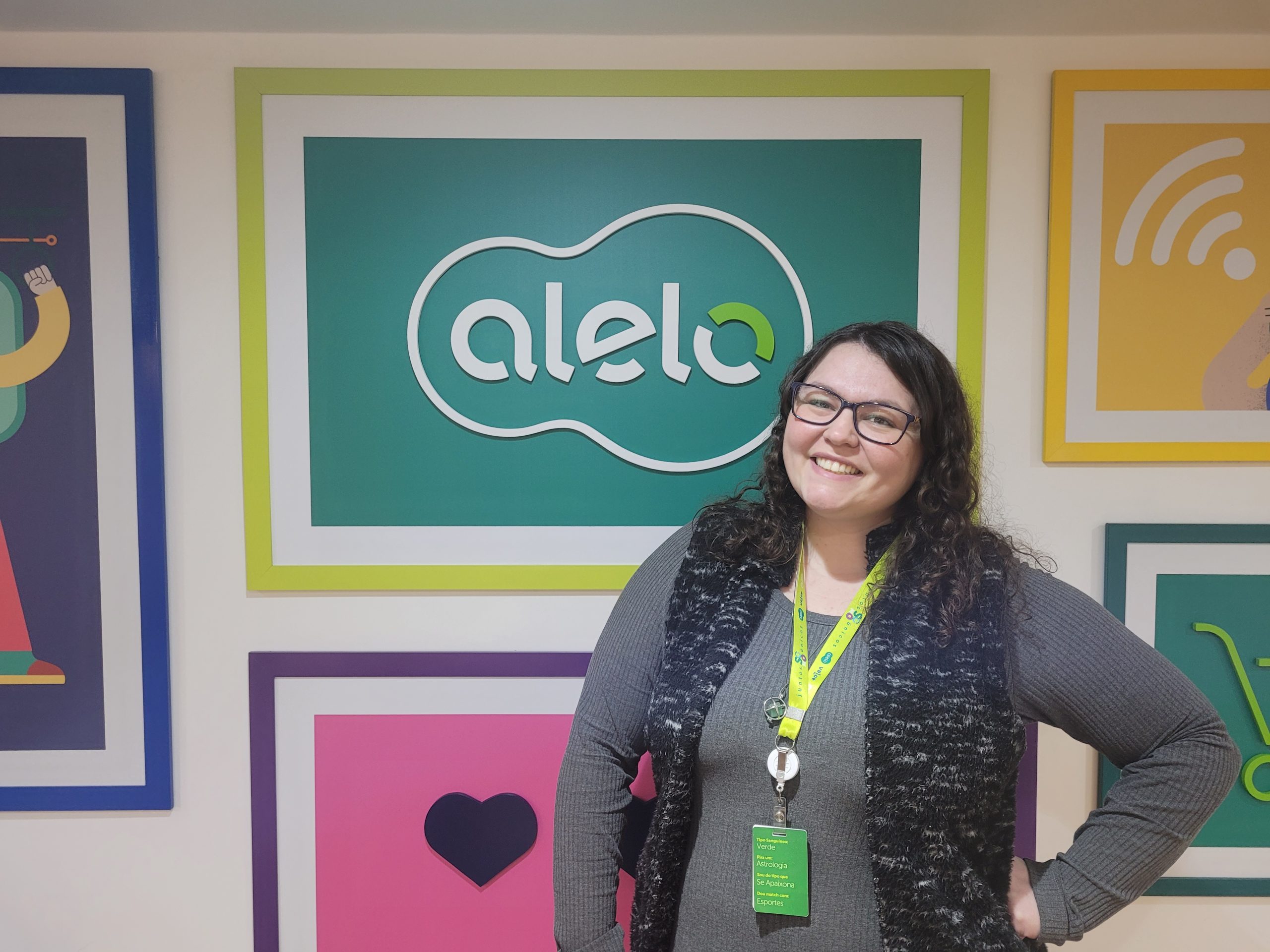 Camila, colaboradora da Alelo fazendo pose em frente a um quadro com o logo da Alelo