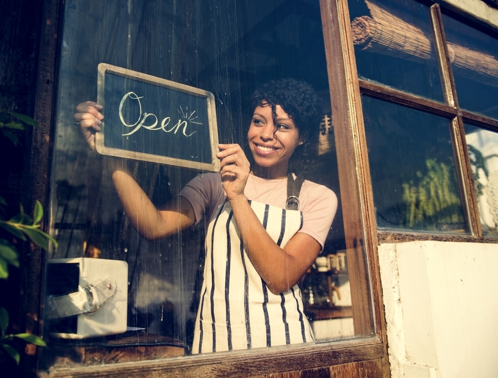 Mulher negra, de avental, sorrindo enquanto segura uma placa escrito "open" na porta de um novo comércio