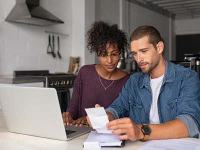 Mulher negra de camisa roxa, ao lado e um homem branco de camisa jeans, olhando as contas em frente a um notebook