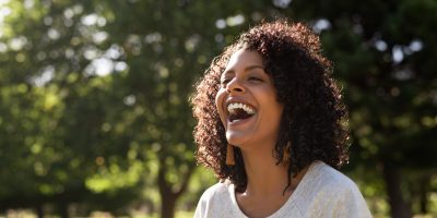 Mulher negra, de cabelo afro, camiseta branca, rindo. Ao fundo um bosque