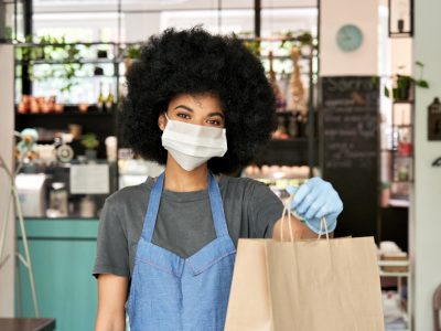 Mulher, com blackpower, vestindo máscara e luvas, um avental, entrega uma sacola de papelão na frente de um estabelecimento comercial