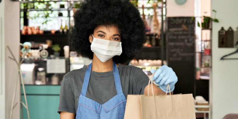 Mulher, com blackpower, vestindo máscara e luvas, um avental, entrega uma sacola de papelão na frente de um estabelecimento comercial