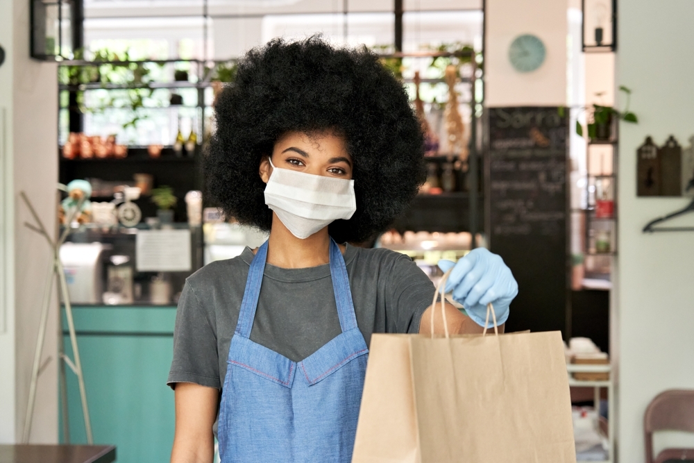 Mulher, com blackpower, vestindo máscara e luvas, um avental, entrega uma sacola de papelão na frente de um estabelecimento comercial