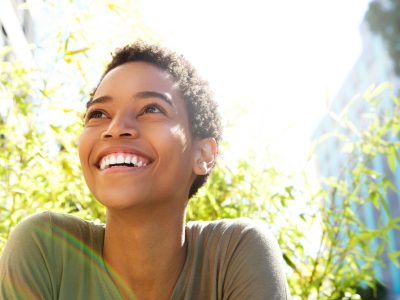 Mulher negra, de cabelos curtos, vestindo blusa cinza, sorrindo num lugar ensolarado