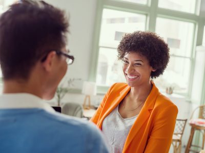 Mulher negra, de terno laranja, sorri para homem de óculos