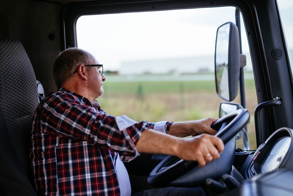 Homem branco, de cabelos grisalhos, veste camisa xadrez, usa óculos e dirige um caminhão