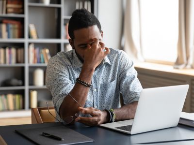 Homem negro, de camisa, sentado em frente ao notebook com um ar de preocupação.