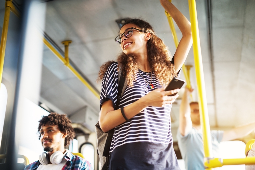 mulher branca, de cabelos cacheados, blusa listrada branco e preto, de óculo, de pé num ônibus vendo seu celular