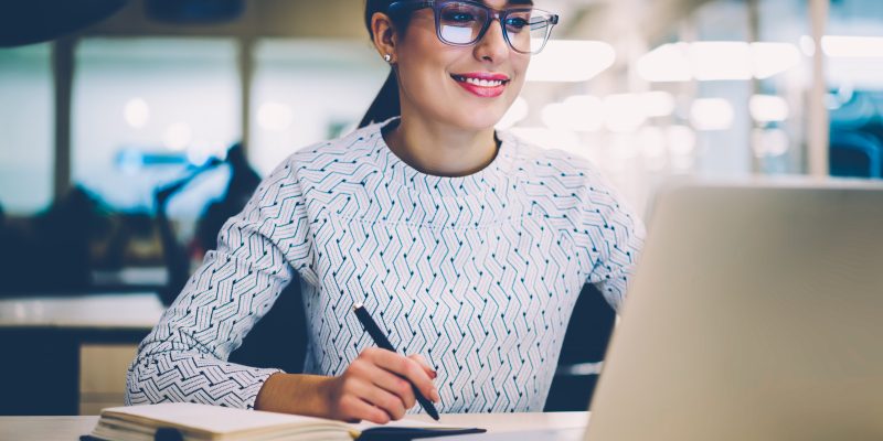 Mulher branca, de óculos, cabelos presos, blusa de cor clara, trabalha em um notebook, segurando uma caneta