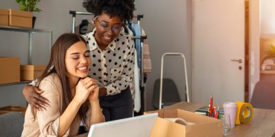 Mulher negra, vestindo camisa branca com estampas pretas, abraça mulher branca sorrindo, enquanto elas olham um notebook na mesa.