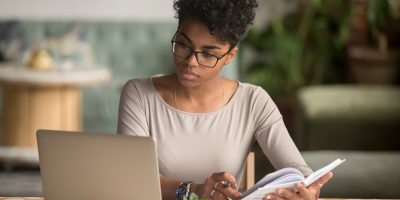 Mulher negra, de cabelos presos, óculos, trabalha em notebook