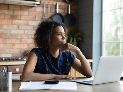 Mulher, negra, com cabelo na altura dos ombros, estilo black power, vestindo blusa de mangas curtas, cor azul marinho, sentada na frente de um notebook com ar pensativo