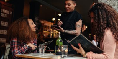 Mulheres olhando um cardápio numa mesa de bar enquanto atendente sorri