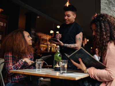 Mulheres olhando um cardápio numa mesa de bar enquanto atendente sorri