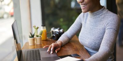 Mulher, preta, com óculos e blusa cinza, sorrindo enquanto trabalha em um notebook em um café