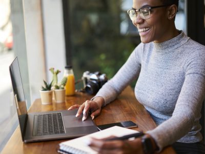 Mulher, preta, com óculos e blusa cinza, sorrindo enquanto trabalha em um notebook em um café