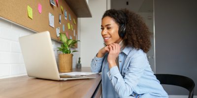 Mulher preta, de cabelos encaracolados, na altura dos ombros, vestindo jaqueta jeans, fones de ouvido, sorrindo enquanto trabalha num notebook