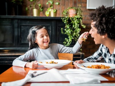 Homem preto, de camisa xadrez, sorri enquanto sua filha, uma criança, branca, de cabelos lisos, vestindo cinza, o alimenta com macarrão.