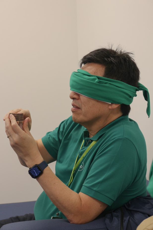Homem vendado, vestindo camisa verde, segura um objeto em suas mãos.
