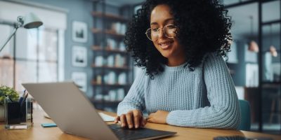 Mulher negra, de blusa azul, óculos, trabalhando em um notebook