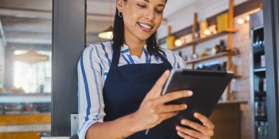 mulher, parda, de avental, sorrindo enquanto lê um tablet em frente ao seu negócio