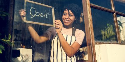 Mulher negra, de avental, sorrindo enquanto segura uma placa escrito "open" na porta de um novo comércio