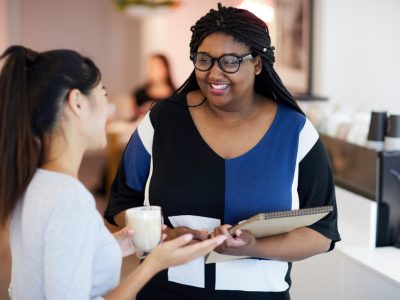 Mulher negra, de óculos, conversa com mulher branca, enquanto sorriem num ambiente de trabalho