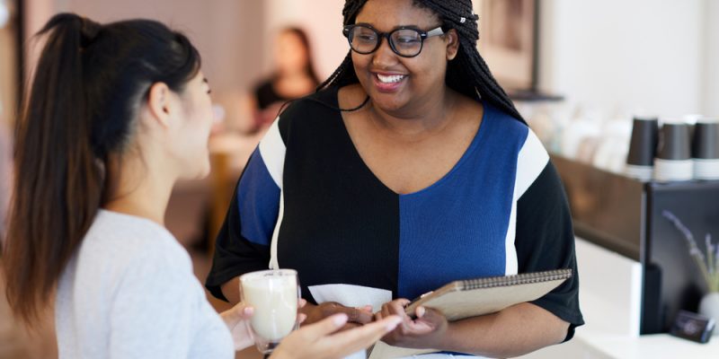 Mulher negra, de óculos, conversa com mulher branca, enquanto sorriem num ambiente de trabalho