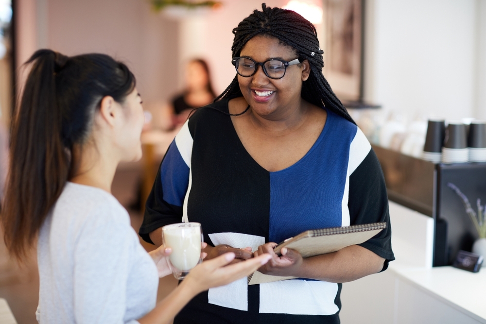 Mulher negra, de óculos, conversa com mulher branca, enquanto sorriem num ambiente de trabalho