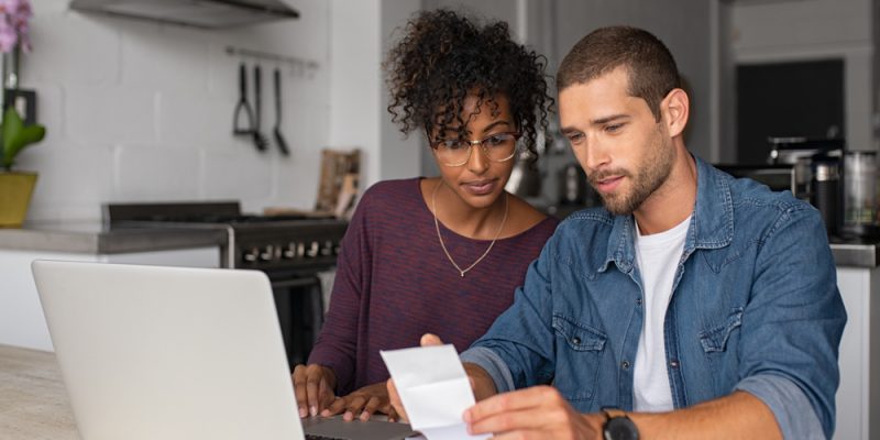 Mulher negra de camisa roxa, ao lado e um homem branco de camisa jeans, olhando as contas em frente a um notebook