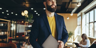 Homem branco, vestindo camiseta amarela e blazer, com notebook na mão