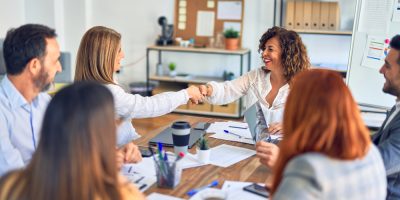 grupo de mulheres profissionais em volta de uma mesa de trabalho, cumprimentando uma delas