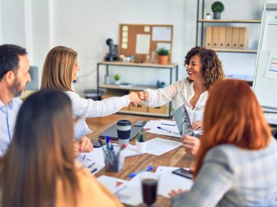 grupo de mulheres profissionais em volta de uma mesa de trabalho, cumprimentando uma delas