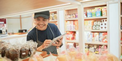 Homem de boné e avental, fazendo anotações enquanto observa uma prateleira cheia de pães