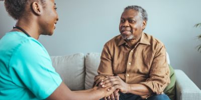 Homem idoso, vestindo camisa marrom segura as mãos de uma médica