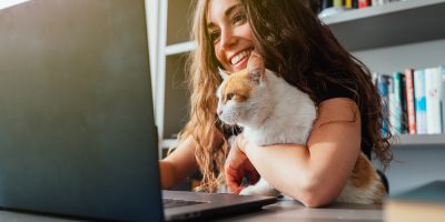 Mulher branca,com cabelos longos e escuros, de blusa preta,segura um gato no colo enquanto trabalha em um notebook