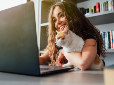 Mulher branca,com cabelos longos e escuros, de blusa preta,segura um gato no colo enquanto trabalha em um notebook