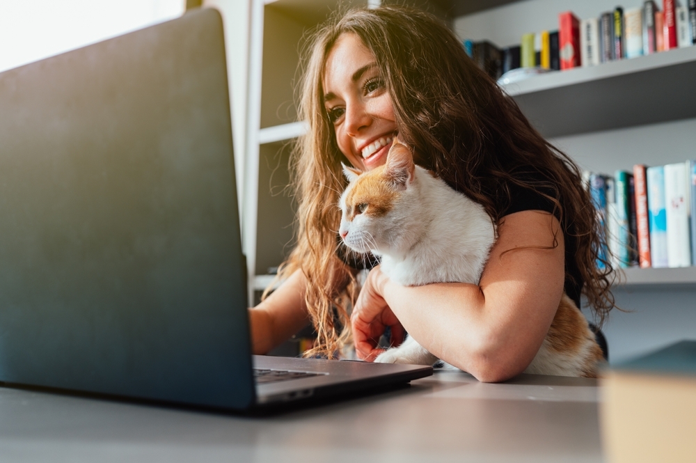 Mulher branca,com cabelos longos e escuros, de blusa preta,segura um gato no colo enquanto trabalha em um notebook