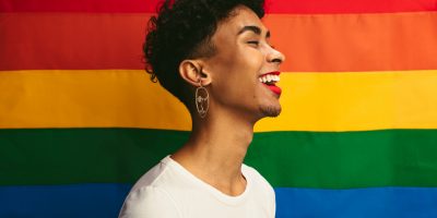 Pessoa de gênero fuído,cabelos curtos, barba, batom e camiseta branca, e frente a uma bandeira colorida LGBTQIA+