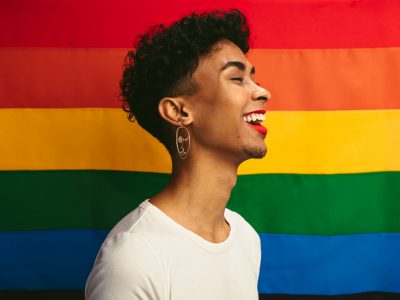 Pessoa de gênero fuído,cabelos curtos, barba, batom e camiseta branca, e frente a uma bandeira colorida LGBTQIA+