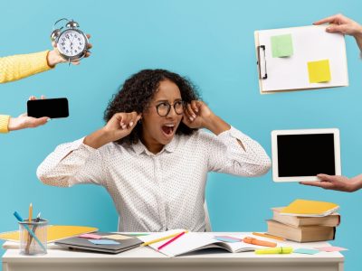 Mulher negra, de cabelos nos ombros, óculos e camisa branca, coloca as mãos nos ouvidos em sinal de estresse ao excesso de trabalho