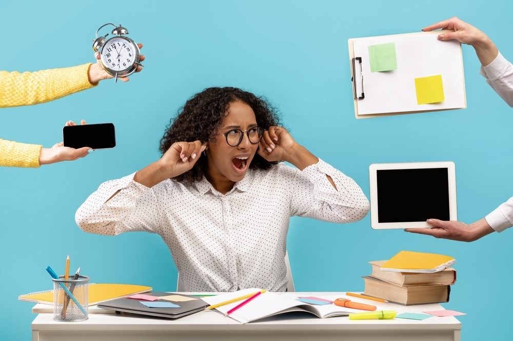 Mulher negra, de cabelos nos ombros, óculos e camisa branca, coloca as mãos nos ouvidos em sinal de estresse ao excesso de trabalho