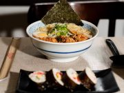 Tigela de ramen sobre mesa forrada com tecido bege, contendo macarrão, ovo cozido, carne empanada, broto de feijão, cebolinha e folha de alga. Em primeiro plano, prato preto com quatro fatias de sushi enrolado em alga, recheado com arroz e legumes. Ao lado, hashi em embalagem de papel e colher preta. Ao fundo, janela desfocada e cadeira escura.