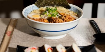 Tigela de ramen sobre mesa forrada com tecido bege, contendo macarrão, ovo cozido, carne empanada, broto de feijão, cebolinha e folha de alga. Em primeiro plano, prato preto com quatro fatias de sushi enrolado em alga, recheado com arroz e legumes. Ao lado, hashi em embalagem de papel e colher preta. Ao fundo, janela desfocada e cadeira escura.