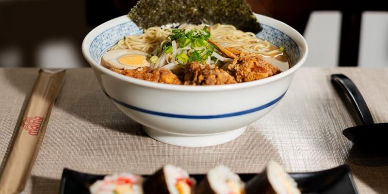 Tigela de ramen sobre mesa forrada com tecido bege, contendo macarrão, ovo cozido, carne empanada, broto de feijão, cebolinha e folha de alga. Em primeiro plano, prato preto com quatro fatias de sushi enrolado em alga, recheado com arroz e legumes. Ao lado, hashi em embalagem de papel e colher preta. Ao fundo, janela desfocada e cadeira escura.