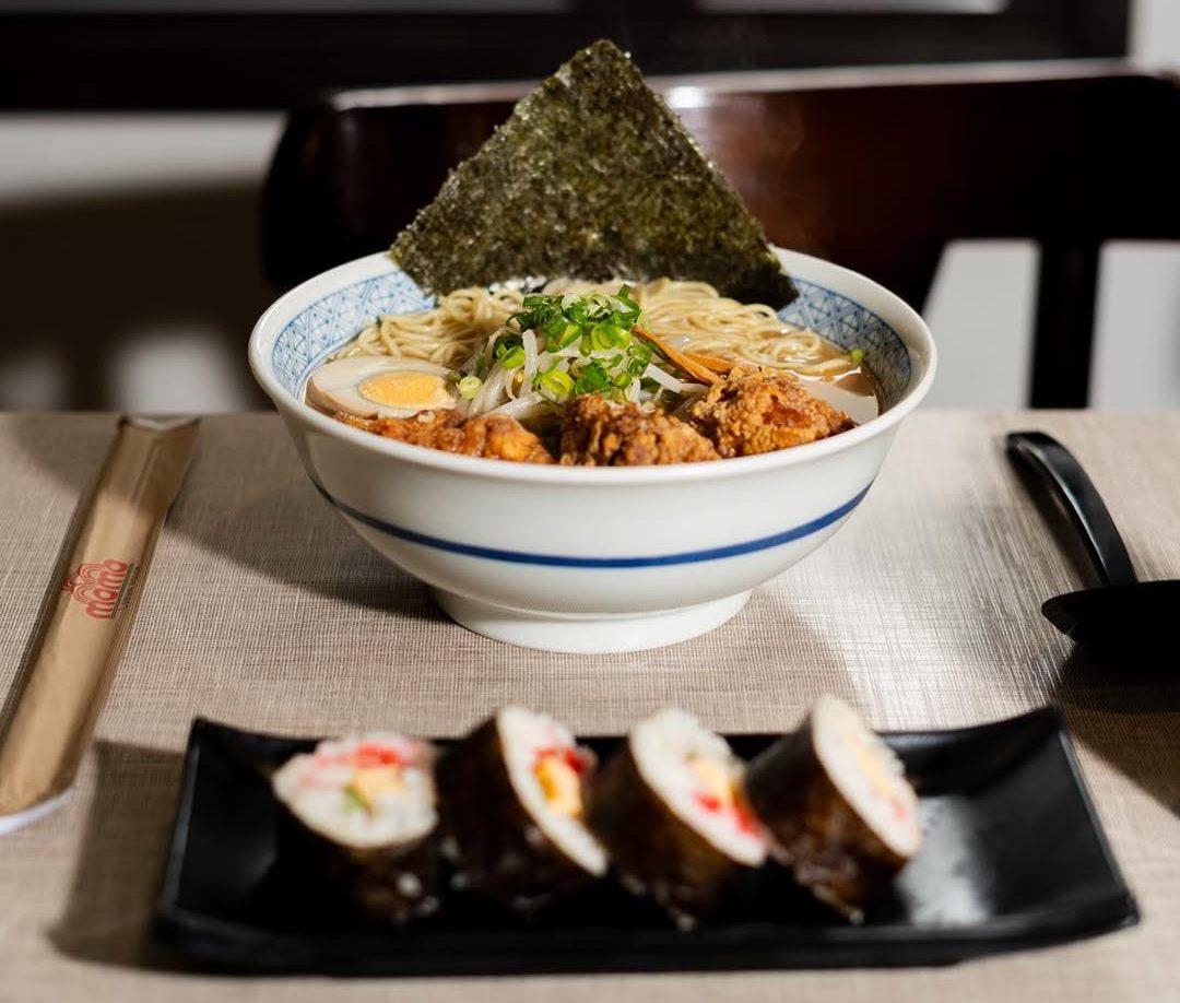 Tigela de ramen sobre mesa forrada com tecido bege, contendo macarrão, ovo cozido, carne empanada, broto de feijão, cebolinha e folha de alga. Em primeiro plano, prato preto com quatro fatias de sushi enrolado em alga, recheado com arroz e legumes. Ao lado, hashi em embalagem de papel e colher preta. Ao fundo, janela desfocada e cadeira escura.